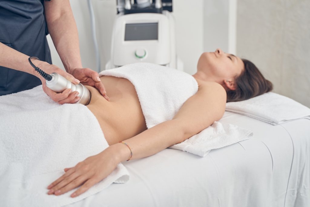 A person lies on a treatment table while a practitioner applies a handheld device to the abdomen in a clinical room.