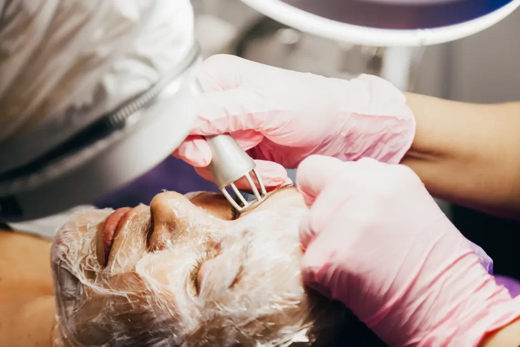 Practitioner wearing pink gloves holds a laser device above a person’s closed eyelid during a facial procedure.